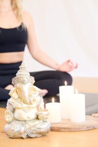Goldene Ganesha-Statue neben brennenden Kerzen, im Hintergrund sitzt eine Yoga Lehrerin aus Leobersdorf in Meditationshaltung.